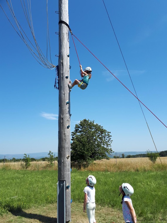 Mädchen klettert gesichert an einem Holzstamm, zwei Kinder schauen zu